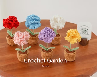Op de afbeelding: Een verzameling van zeven gehaakte anjerbloemen in verschillende kleuren, elk in een bruine gehaakte pot. De bloemen zijn gerangschikt op een houten tafel met een bord waarop staat "Handmade with Love Crochet Garden By Layla".