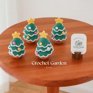 Può includere: Quattro alberi di Natale all'uncinetto con dettagli bianchi e gialli su un tavolo di legno. Gli alberi sono verdi e bianchi con stelle gialle in cima. Il tavolo è marrone e ha un cartello bianco che dice "Handmade with Love" e "Crochet Garden by Layla".