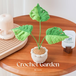 以下が含まれることがあります： 白い鉢に植えられた緑の葉を持つ、かぎ針編みで作られた植物。植物は木製のテーブルの上に置かれ、テーブルには「Crochet Garden by Layla」という文字が書かれている。白いカードには「Made with love Crochet Garden by Layla」という文字が書かれている。