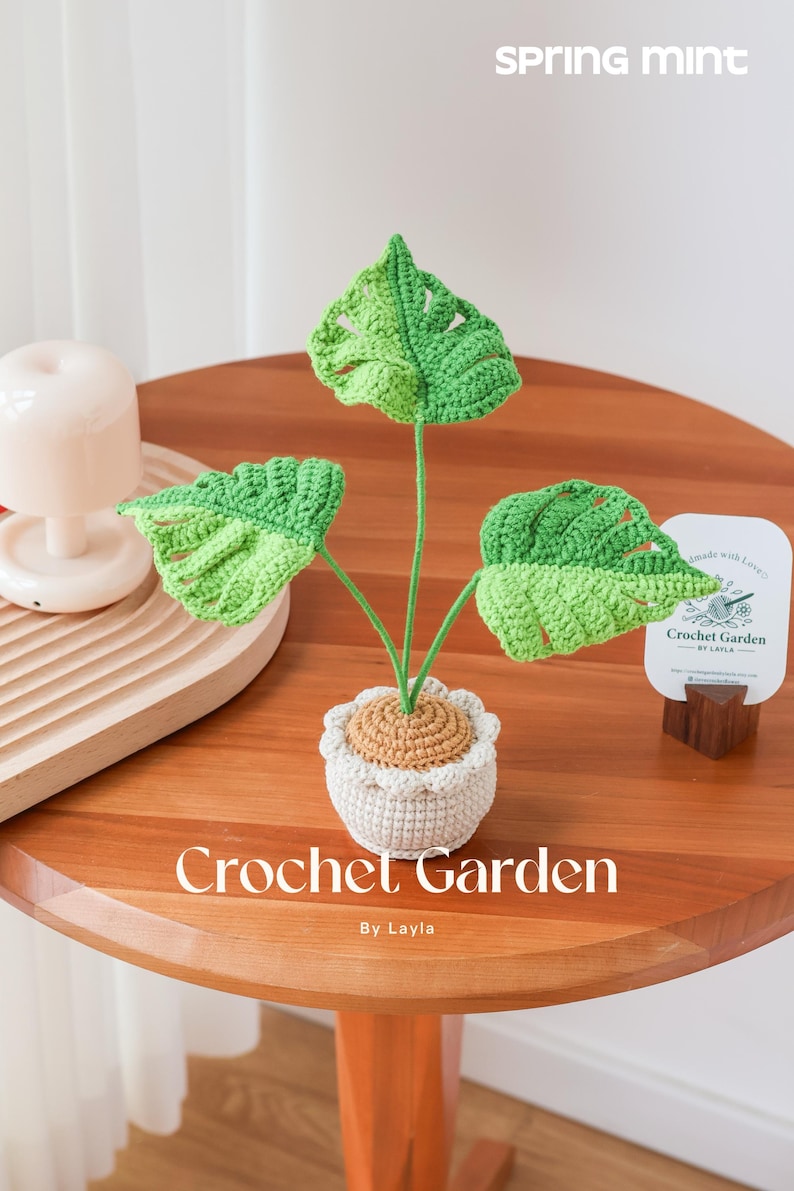 以下が含まれることがあります： 白い鉢に植えられた緑の葉を持つ、かぎ針編みで作られた植物。植物は木製のテーブルの上に置かれ、テーブルには「Crochet Garden by Layla」という文字が書かれている。白いカードには「Made with love Crochet Garden by Layla」という文字が書かれている。
