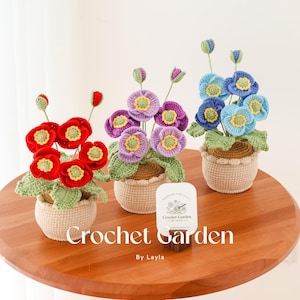 May include: Three crocheted flower arrangements in red, purple, and blue, each in a beige crocheted pot. The pots are sitting on a wooden table. A white card with the text "Crochet Garden by Layla" is in front of the pots.