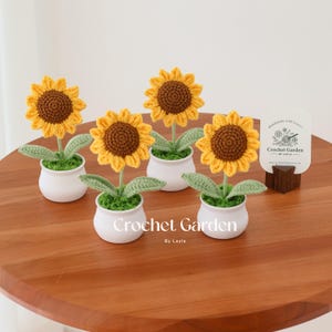 May include: Crocheted sunflowers in white pots with green leaves and brown centers. The sunflowers are arranged on a wooden table. The text "Crochet Garden By Layla" is visible on the front of the pots.