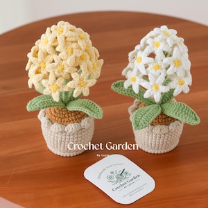 Puede incluir: Dos plantas en maceta de ganchillo con flores blancas y amarillas. Las macetas son de color beige con detalles marrones. Las flores están hechas de hilo y tienen un aspecto realista. El texto "Crochet Garden by Layla" es visible en una tarjeta en primer plano.
