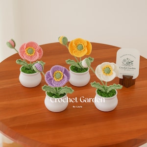 May include: Five crocheted poppy flower arrangements in white, yellow, pink, and purple pots. Each flower is handmade with a green stem and leaves. The pots are white and have a small amount of green moss at the base of the flowers. The flowers are arranged on a wooden table. The text "Crochet Garden by Layla" is visible on the table.