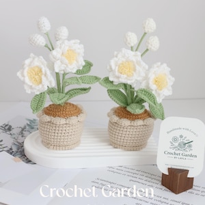 May include: Two crocheted white flower arrangements in beige pots with brown trim. The pots are sitting on a white surface. A business card with the text "Handmade with Love Crochet Garden by Layla" is in the lower right corner of the image.