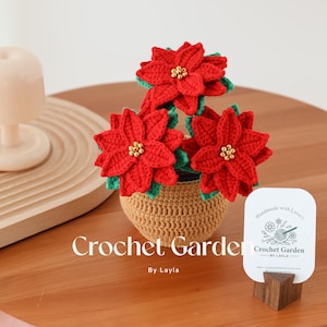 May include: A crocheted poinsettia plant in a brown pot. The plant is made with red and green yarn and has gold beads for the center of the flowers. The pot is sitting on a wooden table. The text "Crochet Garden by Layla" is visible on a white card.