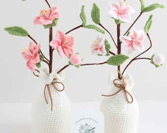 May include: Two crocheted flower arrangements in white ceramic vases. The flowers are pink and white and have green leaves. The vases are tied with brown ribbon. A wooden sign with the text "Handmade with Love Crochet Garden by Layla" is in front of the vases.