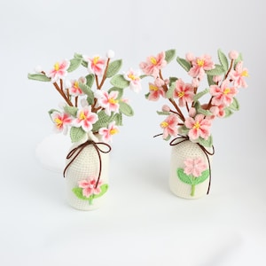 May include: Two crochet flower arrangements in off-white vases. Each vase has a pink flower applique and is tied with a brown string. The flowers are pink and white with yellow centers and green leaves.