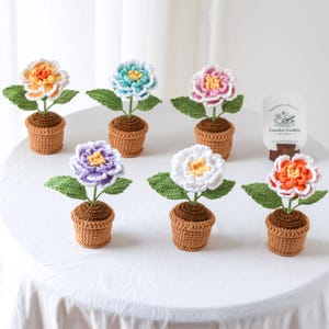 May include: A collection of handmade crochet flowers in small brown pots. Each flower has a different color, including orange, white, purple, pink, and blue. The flowers have green leaves and are arranged on a white surface.