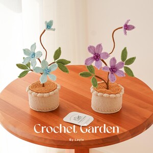 May include: Two crocheted flower arrangements in pots. The flowers are light blue and purple. The pots are beige with a brown crocheted rim. The text "Crochet Garden By Layla" is on a white card in the centre of the table.