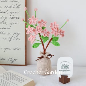 May include: A crocheted bouquet of pink flowers with green leaves in a beige crocheted vase. The vase has a ruffled top and a crocheted design. The flowers are arranged in a vase on a wooden surface. The text "Crochet Garden by Layla" is visible on a white card.