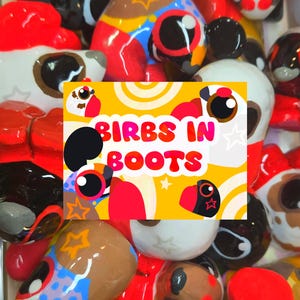 May include: A collection of small, glossy, multi-colored bird figurines featuring various patterns and colors, centered around a rectangular yellow sign with the text BIRBS IN BOOTS in bold red letters, surrounded by stylized star and circle graphics.