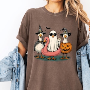 May include: Brown t-shirt featuring a Halloween-themed graphic. The design includes three cartoon geese dressed as a witch, a ghost with sunglasses, and a jack-o'-lantern. A pink flamingo floatie and a bat are also included in the design.