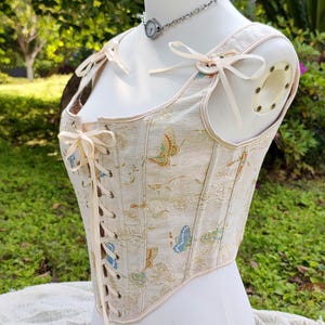 May include: A cream-colored corset with a butterfly pattern in shades of blue, orange, and gold. The corset features cream-colored ribbon ties, shoulder straps with bows, and a delicate trim. The garment is displayed on a mannequin.