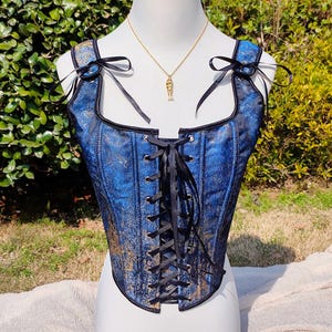 Könnte beinhalten: Ein blau-goldenes Korsett mit schwarzem Besatz und Schnürung vorne. Das Korsett hat schwarze Bandträger und Schleifen an den Schultern. Eine goldene Halskette mit einem Fischanhänger hängt vor dem Korsett. Das Korsett wird auf einer Schaufensterpuppe ausgestellt.