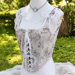 May include: A beige corset with a floral pattern in black and white. The corset has white trim, straps, and a lace-up front. The straps are tied with white bows. A silver necklace with a small watch pendant is also visible.