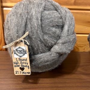 May include: A large ball of natural gray wool roving. A tag tied with twine reads "Fairchild Mercantile" and "1 Pound Nat. Gray Wool Roving 27.7 micron". The wool is a light gray color and appears soft and fluffy, suitable for fiber art projects.