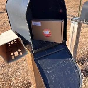 May include: A brown cardboard box with a white label that says "Fairchild Farms" and a red logo with a sun and a field of crops. The box is inside a black mailbox with a black flap that says "Postmaster General Approved By The Postmaster".