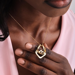 May include: A gold-colored necklace with two pendants. One pendant is a diamond shape with the names "Isabella" and "Camila" engraved. The other pendant is a circle with a mountain design. The necklace is worn on a person.