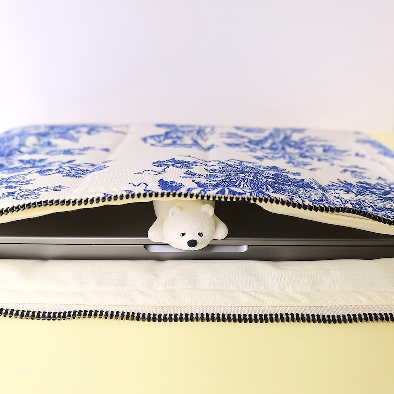 May include: A white laptop case with a blue and white floral pattern. A white polar bear figurine is peeking out from under the case.
