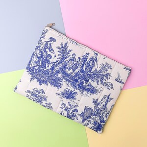 May include: A white zippered pouch with a blue and white toile pattern featuring a scene of people in a garden. The pouch is made of quilted fabric.
