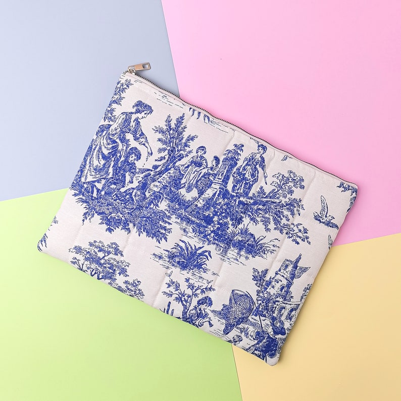 May include: A white zippered pouch with a blue and white toile pattern featuring a scene of people in a garden. The pouch is made of quilted fabric.