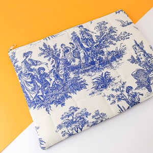 May include: A white clutch bag with a blue and white toile pattern featuring a pastoral scene with figures and trees. The bag has a quilted texture and a gold zipper.