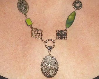 May include: A bronze necklace with an ornate pendant and various decorative elements. The necklace features a mix of shapes, including a leaf, a square, and a circle, with green and yellow accents. The pendant is an oval shape with a detailed filigree design.