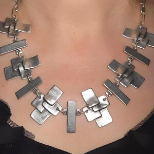 May include: A silver-toned necklace featuring a series of interconnected, overlapping rectangular metal plates. The plates have a brushed, matte finish and are linked together by small metal rings, forming a wide, structured collar design.