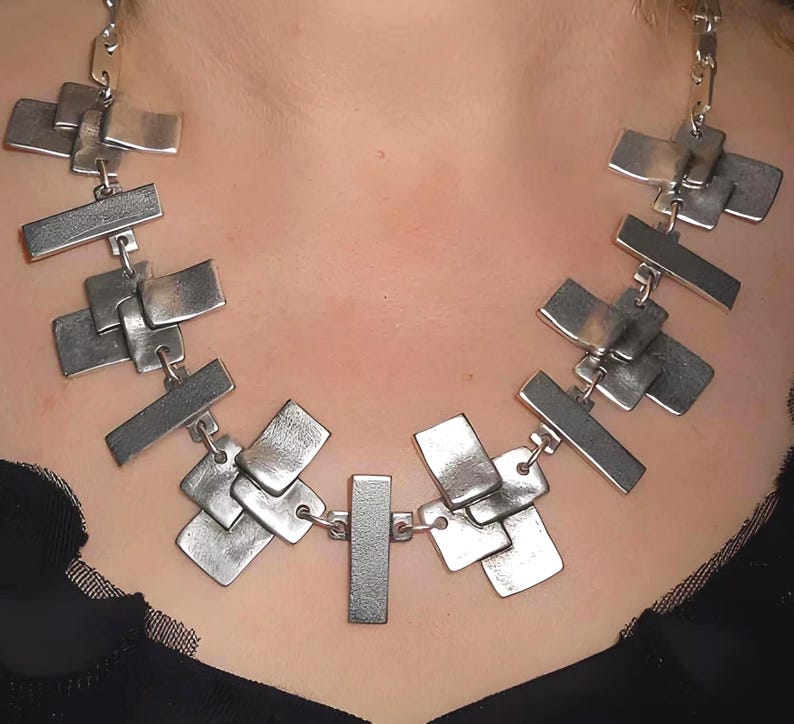 May include: A silver-toned necklace featuring a series of interconnected, overlapping rectangular metal plates. The plates have a brushed, matte finish and are linked together by small metal rings, forming a wide, structured collar design.