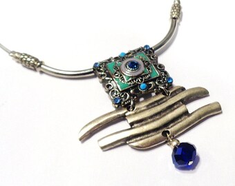 May include: A silver-toned necklace featuring a rigid collar and a central pendant. The pendant includes a square filigree frame with teal enamel, small blue rhinestones, and a dangling dark blue faceted crystal bead at the bottom.
