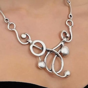 May include: A silver-toned metal necklace featuring an asymmetrical design of interconnected circular loops and smooth, rounded spheres. The chain consists of curved links that transition into a central focal point of overlapping rings and spheres.