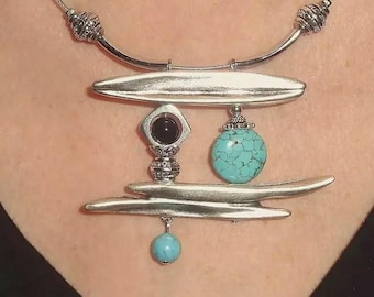 May include: A silver-tone necklace featuring a rigid curved collar with decorative beads. The pendant consists of two horizontal silver bars, a central black stone in a diamond-shaped setting, a round turquoise-coloured bead, and a small dangling blue bead.