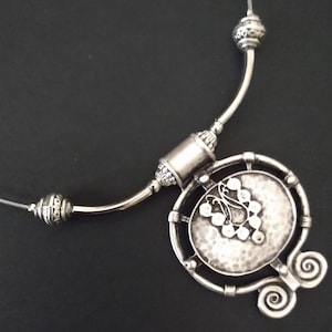 May include: A silver-toned necklace featuring a rigid collar section with two decorative beads and a central circular pendant. The pendant has a hammered texture, a raised butterfly-shaped design, and two spiral accents at the bottom edge.