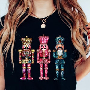 May include: Black t-shirt featuring three faux sequin nutcracker designs. The nutcrackers are in pink, blue, and multicolored sequin patterns. The text on the shirt reads: "The design in the image is Faux Sequin. It is not real sequin."