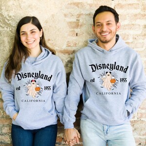 Puede incluir: Dos personas con sudaderas con capucha de Disneyland a juego de color gris claro. Las sudaderas con capucha presentan un fantasma sosteniendo una calabaza, con el texto "Disneyland est 1955 CALIFORNIA". Las sudaderas tienen un bolsillo delantero y una capucha.