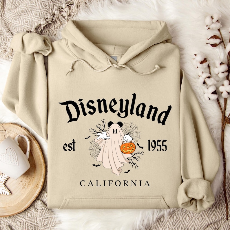 Puede incluir: Sudadera con capucha beige con un dise&ntilde;o de Halloween de Disneyland. El gr&aacute;fico incluye un fantasma sosteniendo una calabaza, telara&ntilde;as, murci&eacute;lagos y el texto "Disneyland est 1955 California."