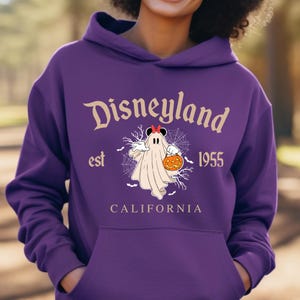 Puede incluir: Una sudadera con capucha morada de Disneyland con un dise&ntilde;o de Halloween. El dise&ntilde;o presenta un fantasma sosteniendo una calabaza, con las palabras "Disneyland est 1955 California" en una fuente vintage.