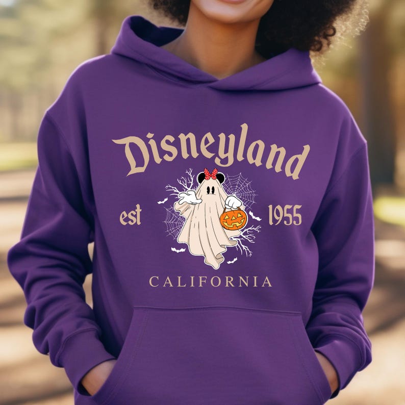 Puede incluir: Una sudadera con capucha morada de Disneyland con un dise&ntilde;o de Halloween. El dise&ntilde;o presenta un fantasma sosteniendo una calabaza, con las palabras "Disneyland est 1955 California" en una fuente vintage.