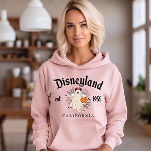 Puede incluir: Una sudadera con capucha rosa claro con un bolsillo delantero y la palabra "Disneyland" en escritura negra. Debajo, un dibujo de un fantasma sosteniendo una calabaza, con el texto "est 1955 CALIFORNIA".