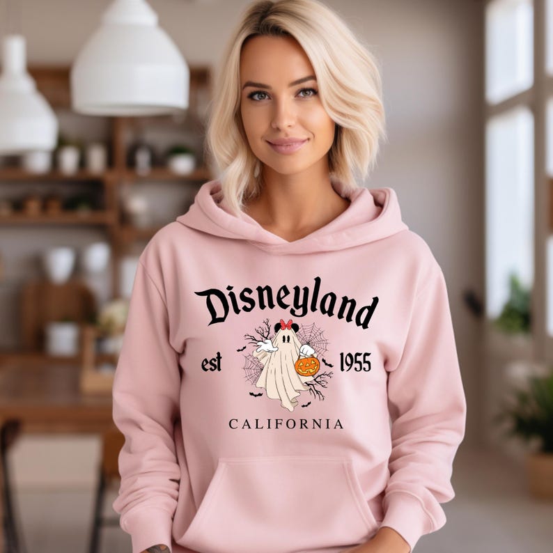 Puede incluir: Una sudadera con capucha rosa claro con un bolsillo delantero y la palabra "Disneyland" en escritura negra. Debajo, un dibujo de un fantasma sosteniendo una calabaza, con el texto "est 1955 CALIFORNIA".