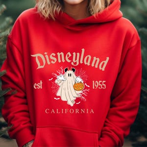 Puede incluir: Una sudadera con capucha roja con el logotipo de Disneyland y un gr&aacute;fico de Halloween. El gr&aacute;fico muestra un fantasma disfrazado de Mickey Mouse sosteniendo una calabaza, con el texto "Disneyland est 1955 California".