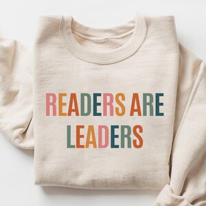 Könnte beinhalten: Ein beiger Pullover mit dem Aufdruck "READERS ARE LEADERS" in bunten Buchstaben. Die Buchstaben sind in den Farben Rosa, Orange, Gelb, Grün, Blau und Braun gehalten.