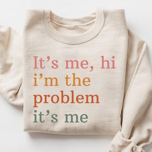 以下が含まれることがあります： ベージュのトレーナーに「It's me, hi i'm the problem it's me」という文字がピンク、オレンジ、赤、緑でプリントされています。