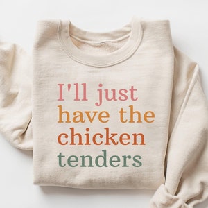 以下が含まれることがあります： レインボーカラーで「I'll just have the chicken tenders」とプリントされたベージュのスウェットシャツです。