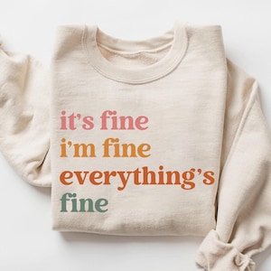 以下が含まれることがあります： レインボーカラーで「it's fine i'm fine everything's fine」とプリントされたベージュのトレーナーです。