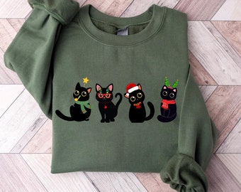May include: A military green sweatshirt with a Christmas-themed design of four black cats. Each cat is decorated with festive accessories such as a star, glasses, a Santa hat, and a Christmas tree headband. The sweatshirt has a crew neck and long sleeves.