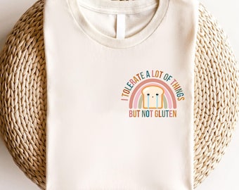 May include: Cream-colored t-shirt with a round neck. The shirt features a graphic of a crying slice of bread with a rainbow and the text "I TOLERATE A LOT OF THINGS BUT NOT GLUTEN". The shirt is displayed on a woven surface.