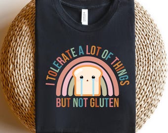 May include: Black t-shirt with a graphic of a crying slice of bread inside a rainbow. Text reads "I TOLERATE A LOT OF THINGS BUT NOT GLUTEN". The shirt is displayed on a woven, circular surface.