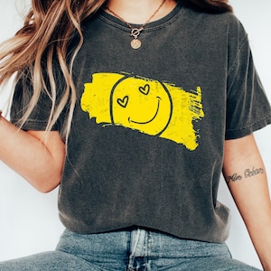 Może przedstawiać: Szary t-shirt z żółtą buźką uśmiechniętą z serduszkami w oczach. Uśmiechnięta buźka znajduje się na żółtym pociągnięciu pędzla.